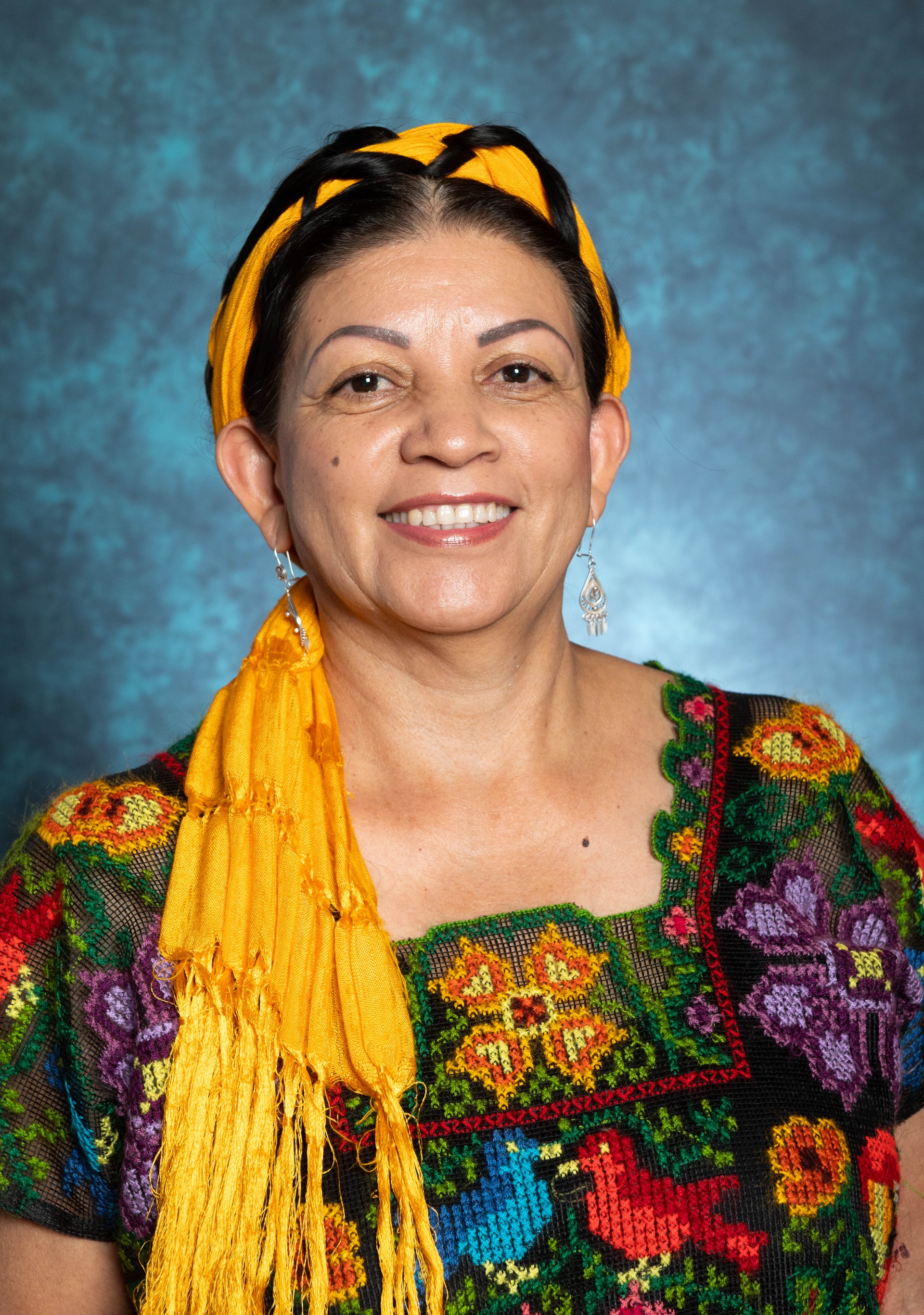 Professional photo of a smiling Claudia Gonzalez Astorga wearing a colorful handmade cross-stich huipil, a vibrant yellow Mexican shawl braided in her long hair over her head, and wearing two small hand-woven silver earrings. Claudia is Mexican, have light brown skin, dark hair, brown eyes, and brown eyebrows.