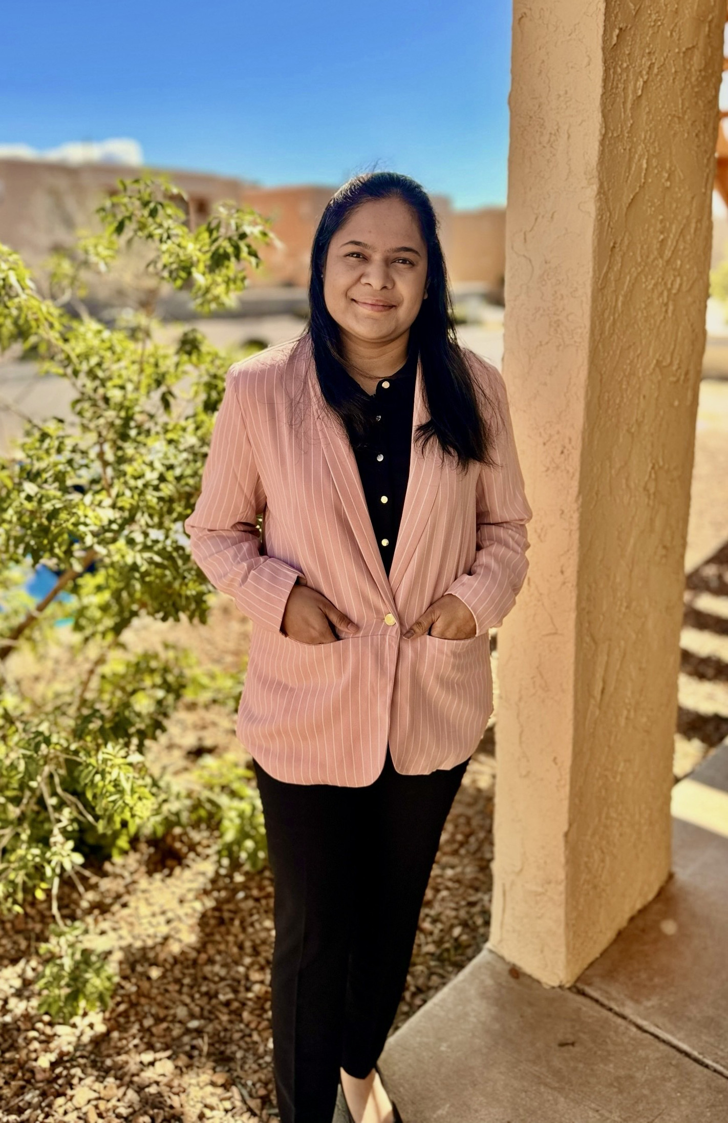 An image of the graduate assistant of Latin American programs, Pracheta Harlikar. The image features a woman standing outdoors against a backdrop of a clear blue sky and bright sunshine. She is positioned next to a beige, textured column of a building, with a green bush and some buildings slightly blurred in the background. She is wearing a light pink pinstripe blazer over a black shirt with buttons and black pants. Her hands are casually tucked into the pockets of her blazer, and she is smiling softly at the camera. The ground around her consists of small rocks and some sparse greenery.