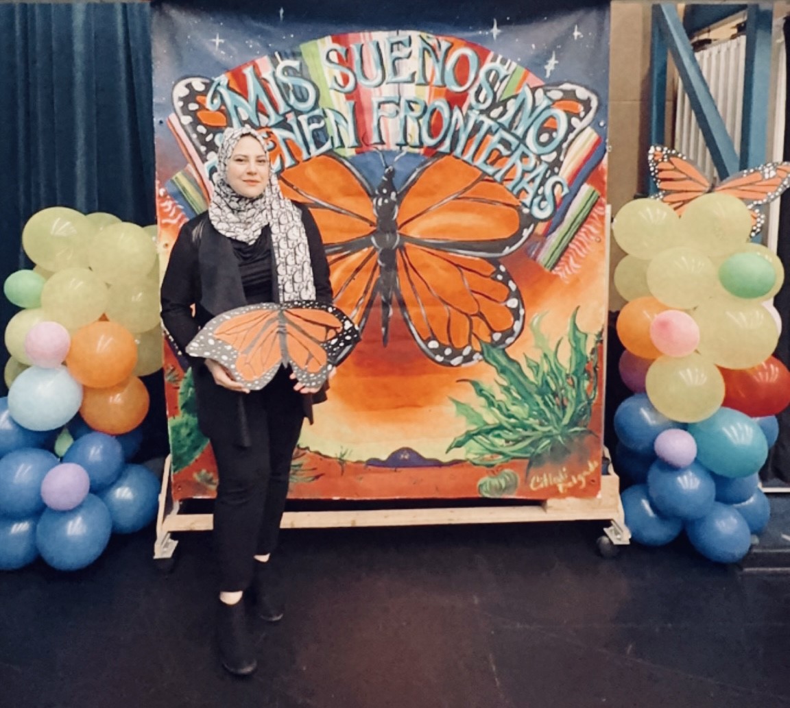 Photo of Jawaher Banihani Graduate Assistant of Latin American Programs. The image features a person standing in front of a colorful, hand-painted backdrop. The backdrop depicts an enormous monarch butterfly with outstretched wings, set against a vibrant background of a sunrise or sunset with a gradient sky transitioning from blue at the top to orange at the bottom. Above the butterfly, bold, colorful text reads "MIS SUEÑOS NO TIENEN FRONTERAS." The person standing in front is holding a large, detailed cutout of a monarch butterfly. They are dressed in dark clothing and have a headscarf with a white and black pattern.