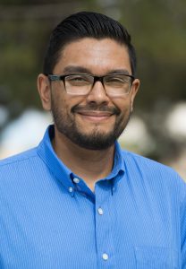 Image of Joseph Duran Administrative Assistant, Intermediate. The image portrays a man from the chest up, standing outdoors. He is wearing a bright blue, button-up shirt with the collar open and glasses with dark, thick frames. He has short, neatly combed black hair and a short, well-groomed beard and mustache. His expression is calm, with a slight smile. The background is blurred with hues of green and white, suggesting greenery or an outdoor setting.