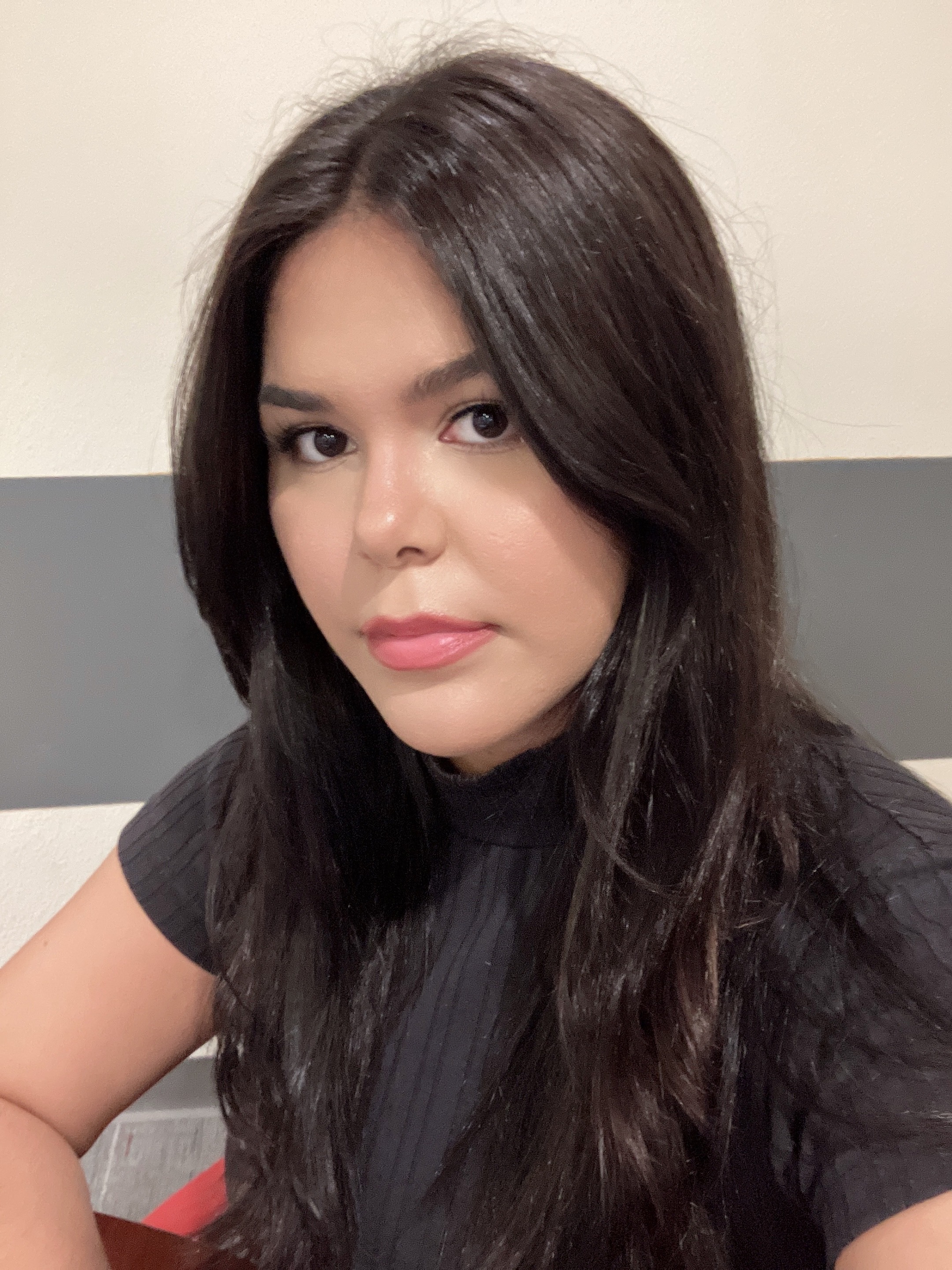 Photo of Destiny Hallett Work-Study for Latin American Programs. The image features a young woman with long, straight, dark brown hair. She is positioned in a three-quarter view, facing the camera with a neutral expression. The woman is dressed in a black, short-sleeved top with a ribbed texture. The background consists of a two-toned wall with the upper portion painted white and a horizontal gray stripe in the middle. The lighting is soft and evenly illuminates her face.