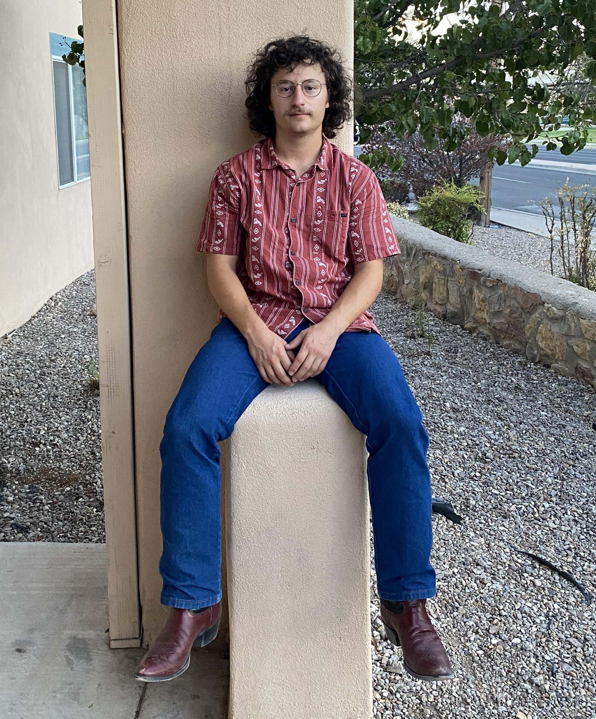 Photo of John Leo Kirkpatrick Work-Study for Latin American Programs. A person is pictured seated on a rectangular, beige column located on an outdoor porch. They are wearing a red, patterned short-sleeve button-up shirt and blue jeans, paired with brown boots. The individual has curly hair, wears glasses, and has a mustache. Their hands are resting on their lap, and they are holding a smartphone with a case in their left hand. The backdrop includes a beige wall, part of a window with a blue frame to the left, a stone ground, and a stone wall to the right. Behind the stone wall, there are green trees and a road in the distance.