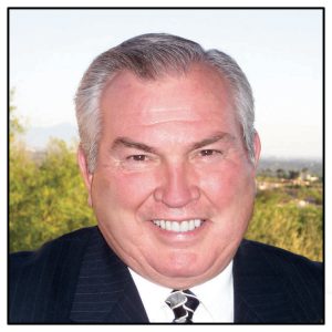 Photo of Dr. Danny Villanueva. The image shows a close-up portrait of an older man smiling directly at the camera. He has short, neatly combed gray hair and is wearing a dark suit jacket over a white dress shirt, complemented by a patterned tie. The background features an outdoor setting with lush greenery and a hazy, distant view of a landscape, possibly including trees and some building structures.