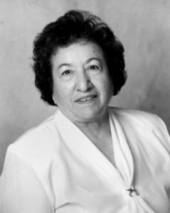 Photo of Dr. Narcisa Zarate. The image is a black and white portrait of an older woman with short, curly dark hair. She is facing the camera and smiling gently. She wears a light-colored blouse with a V-neckline. The background is plain and out of focus, highlighting the subject.