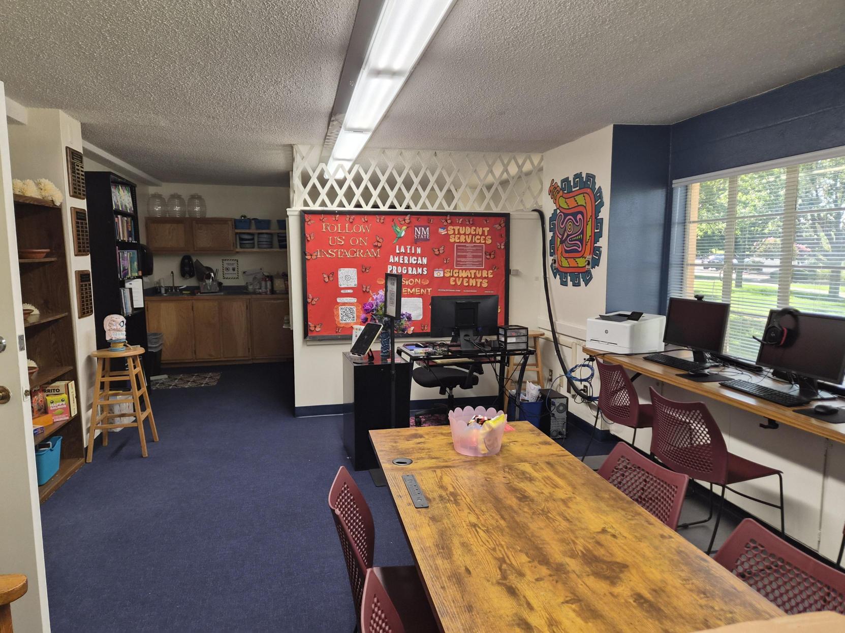 A picture of an the Lab of Latin American Program. The image shows a cozy room featuring a vibrant orange-red couch positioned in front of a brightly colored bulletin board. The board has a green border, with a pink and orange checkered background, and is adorned with numerous photos and captions under the title "SNAPSHOTS OF CHICANX LIFE." The couch has two patterned cushions, one on each side, and a dark green blanket draped over the right armrest. Behind the couch, to the left, there is a small kitchenette area with wooden cabinets, a black refrigerator, and a countertop with various items. To the right of the bulletin board, part of a white wall with a wooden chair rail and decorative wall features is visible.