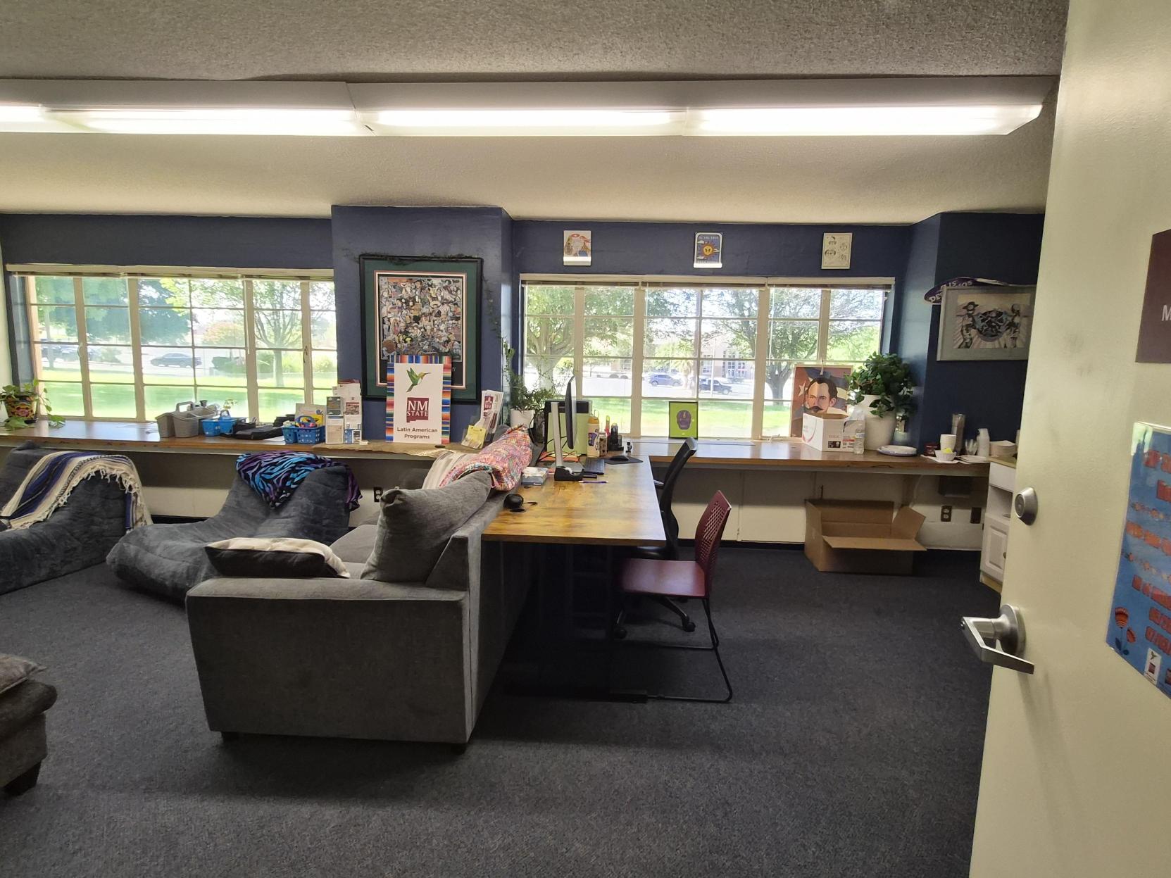 A picture of the study room of Latin American Program. The image depicts a study or meeting room with various decorations and furnishings. The room has a dark carpeted floor and off-white walls. There are two rectangular beige tables placed in the center, surrounded by multiple cushioned chairs in different colors (blue, gray, and black).  In the left corner of the room, a long wooden counter runs against the wall below a wide window with blinds partially open, showing trees outside. On the counter, there is a red coffee machine, snacks, and a variety of supplies including face masks in boxes, a tissue box, and a sign. Colorful paper banners are strung across the top of the window.  On the right side, a black bookshelf filled with books and decorations leans against the wall. Next to the bookshelf stands a large figure of a woman in a purple dress, likely a piñata, with butterfly wings behind it. Several posters and colorful signs with text and images are attached to the walls and surfaces throughout the room.  Text Section:  "Hispanic Council" (on the wall above the paper banners) "8M Maestras Luchando" (on a poster on the counter) "Merezco a mis hijas volver a ver" (on a poster) "Maybe Life" (on a piece of art on the wall) "CA QUE MUDAR DE TU VIDA TENDRIAS QUE MATAR PARA QUE TE IMPORTE VIOLENCIA DE GENERO" (on a poster behind the large figure)