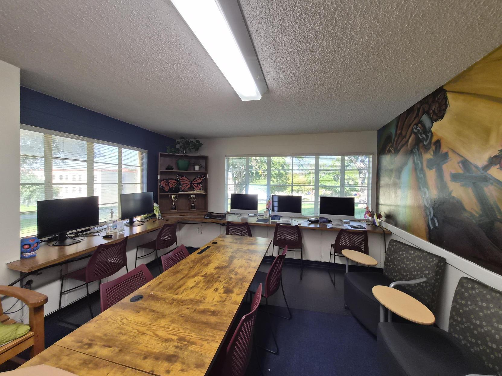 A picture of an the Lab of Latin American Program. The image depicts a mural painted on the wall above a row of desks in a room with multiple computer workstations. The mural is vibrant and spans the entire upper section of the wall, depicting a series of scenes that appear to convey themes of struggle and liberation. The mural is predominantly in shades of yellow, orange, blue, and red colors. The left side shows a muscular figure breaking free from chains, with intense facial expressions and vibrant colors highlighting the action. Towards the center, a figure in a red jacket and blue pants kneels on the ground as if in a moment of effort or reflection, with a large, red, circular emblem featuring a black silhouette of an eagle behind it. On the right, a figure draped in a white and orange garment stands with a solemn expression. The mural is signed in the bottom right corner by "H. Telles & R. Lavadie." Below the mural, there are four workstations with computer monitors, keyboards, and chairs. To the left, there's a window with a curtain partially drawn up and a trail of green hanging plants on the windowsill and wall.