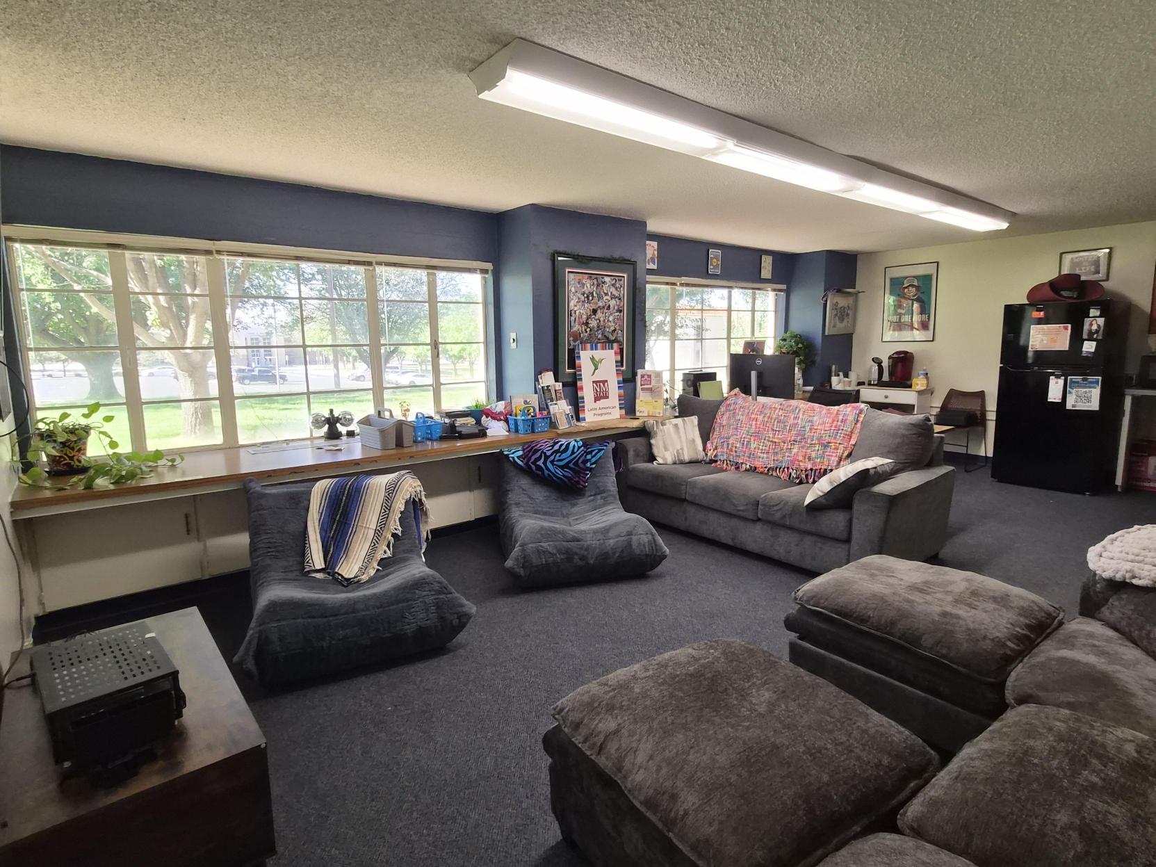 A picture of an the Lab of Latin American Program. The image showcases a small office or classroom space. The room contains two off-white rectangular tables pushed together, surrounded by several chairs with light blue and dark gray fabric upholstery. Against the wall to the right is a large flat-screen TV on a black stand. To the left, there is a whiteboard filled with colorful drawings and text, including hearts, flowers, a strawberry, and welcoming messages. Below the whiteboard, there is a black table with various items like hand sanitizer, bins, flyers, and artwork, including a prominent painting of a pair of red lips. The room has a blue carpeted floor and white walls. In the back left corner, there are a few more chairs with attached writing surfaces stacked near the table.