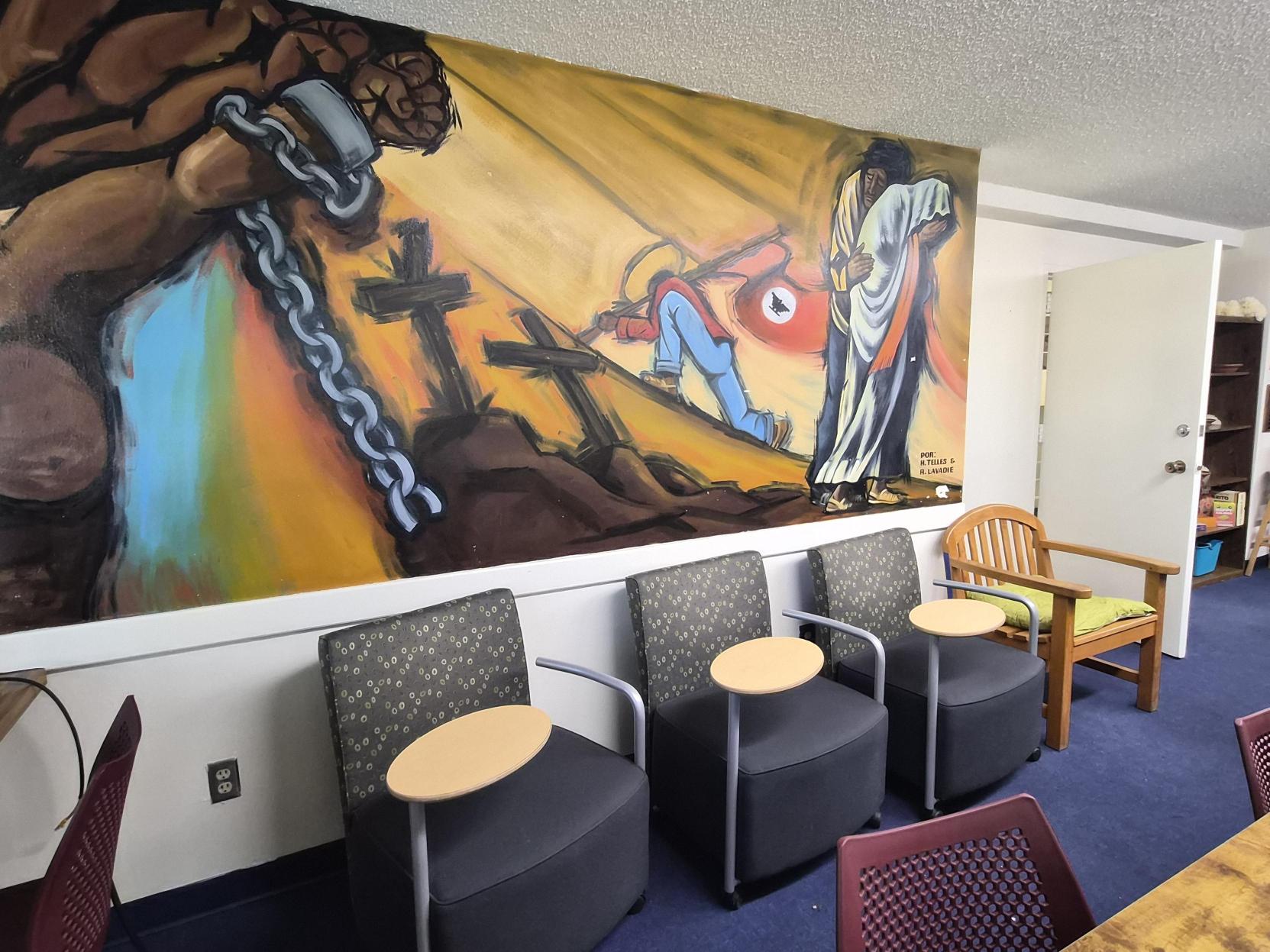 A picture of an the Lab of Latin American Program. The image depicts a small, cozy room that appears to be a workspace or a lounge area. In the foreground on the left, there's a plush red armchair. Behind the armchair and to the right, there’s a corner with a desktop computer setup featuring several desktop computers on a continuous wooden desk against the wall. The desk has three purple office chairs. A poster is mounted above the desk with a colorful and abstract design featuring geometrical patterns and shapes.  On the wall behind the red armchair, two handmade posters rest on the floor against the wall. One has a red section with a figure and the text in black, and the other is white with green handprints and text.  Two windows with blinds allow natural light to filter in, illuminating the room. A large, leafy potted plant in a white pot sits on the desk. On the right side of the image, another handmade poster with pink background text is propped against the wall with illustrations of cartoon characters.
