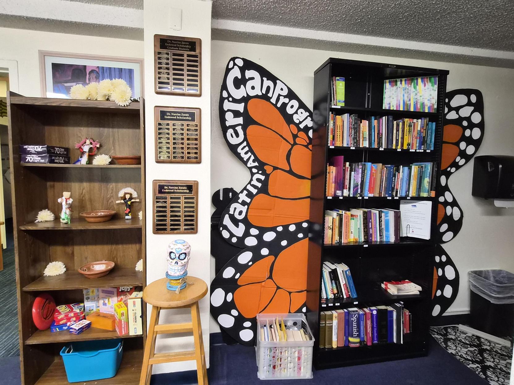 A picture of an the Lab of Latin American Program. The image shows a corridor leading to a room labeled "Chicano Programs Lounge." On the left side of the corridor is a large bulletin board covered with a light blue background and bordered in red with multiple colorful flyers and announcements pinned to it. Above the entry door to the lounge, there is a dark blue sign with white lettering that reads "Chicano Programs Lounge." The door has a whiteboard mounted on it with handwritten red text. Through the open door, an interior view of the lounge is visible, showing a red couch with several pillows in front of a wall decorated with various posters and art. Beside the door, on the right wall, there is a large, vibrant mural featuring a stylized Aztec or Mayan figure with green skin, a yellow and orange headdress, and crossed arms, above the word "LOUNGE" in green letters.
