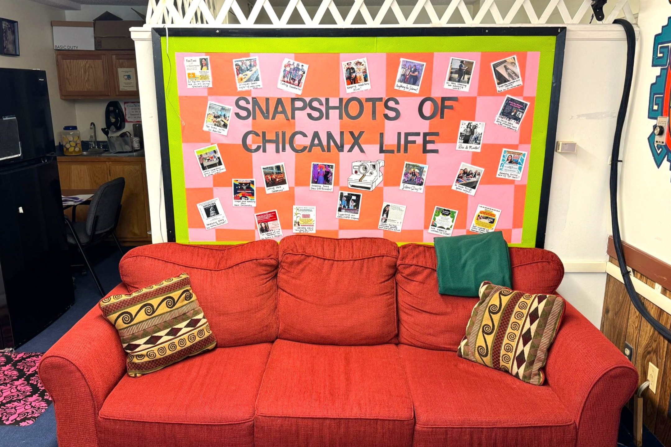 A picture of an the Lab of Latin American Program. The image shows a cozy room featuring a vibrant orange-red couch positioned in front of a brightly colored bulletin board. The board has a green border, with a pink and orange checkered background, and is adorned with numerous photos and captions under the title "SNAPSHOTS OF CHICANX LIFE." The couch has two patterned cushions, one on each side, and a dark green blanket draped over the right armrest. Behind the couch, to the left, there is a small kitchenette area with wooden cabinets, a black refrigerator, and a countertop with various items. To the right of the bulletin board, part of a white wall with a wooden chair rail and decorative wall features is visible.