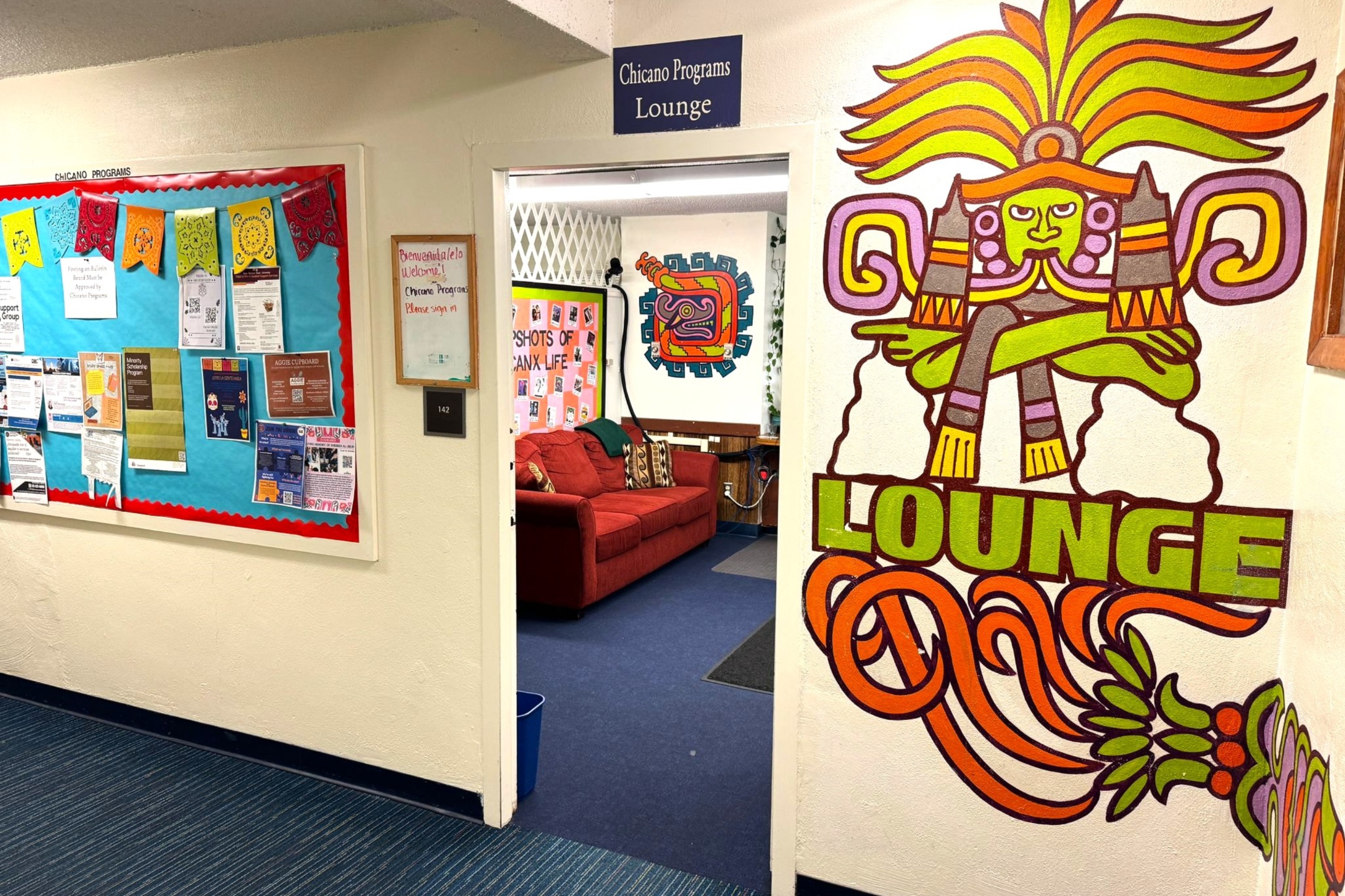 A picture of an the Lab of Latin American Program. The image shows a corridor leading to a room labeled "Chicano Programs Lounge." On the left side of the corridor is a large bulletin board covered with a light blue background and bordered in red with multiple colorful flyers and announcements pinned to it. Above the entry door to the lounge, there is a dark blue sign with white lettering that reads "Chicano Programs Lounge." The door has a whiteboard mounted on it with handwritten red text. Through the open door, an interior view of the lounge is visible, showing a red couch with several pillows in front of a wall decorated with various posters and art. Beside the door, on the right wall, there is a large, vibrant mural featuring a stylized Aztec or Mayan figure with green skin, a yellow and orange headdress, and crossed arms, above the word "LOUNGE" in green letters.