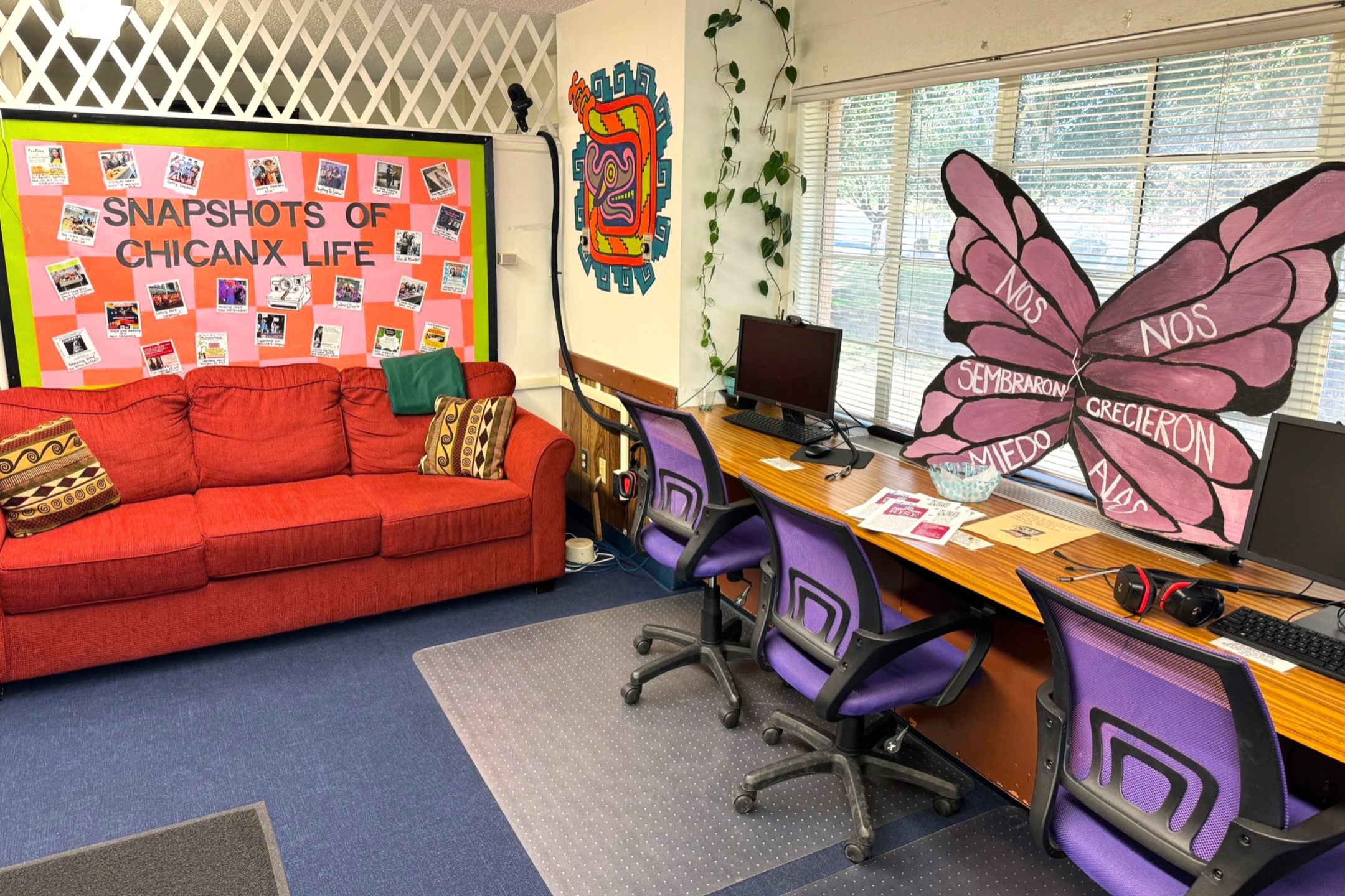 A picture of an the Lab of Latin American Program. The image depicts a vibrant, welcoming room with a combination of seating and workspace. On the left side of the image is a red fabric couch adorned with patterned cushions. Above the couch, a large bulletin board is mounted on the wall with a green border and a central pink area. The bulletin board is titled "SNAPSHOTS OF CHICANX LIFE" and is decorated with numerous small photos and printed materials.  To the right, a wooden desk spans the length of the wall below a series of windows with horizontal blinds. The desk has multiple workstations equipped with computer monitors, keyboards, and purple mesh office chairs. On the corner of the desk closest to the couch is a large butterfly cutout with pink wings, inscribed with the words "NOS SEMBRARON MIEDO," "NOS CRECIERON ALAS." Above the desk area, the wall is adorned with a brightly colored mural featuring abstract, geometric shapes surrounding a stylized face, complemented by climbing plants on the wall.  The floor consists of a combination of blue carpet and a clear, studded plastic mat under the desk chairs.