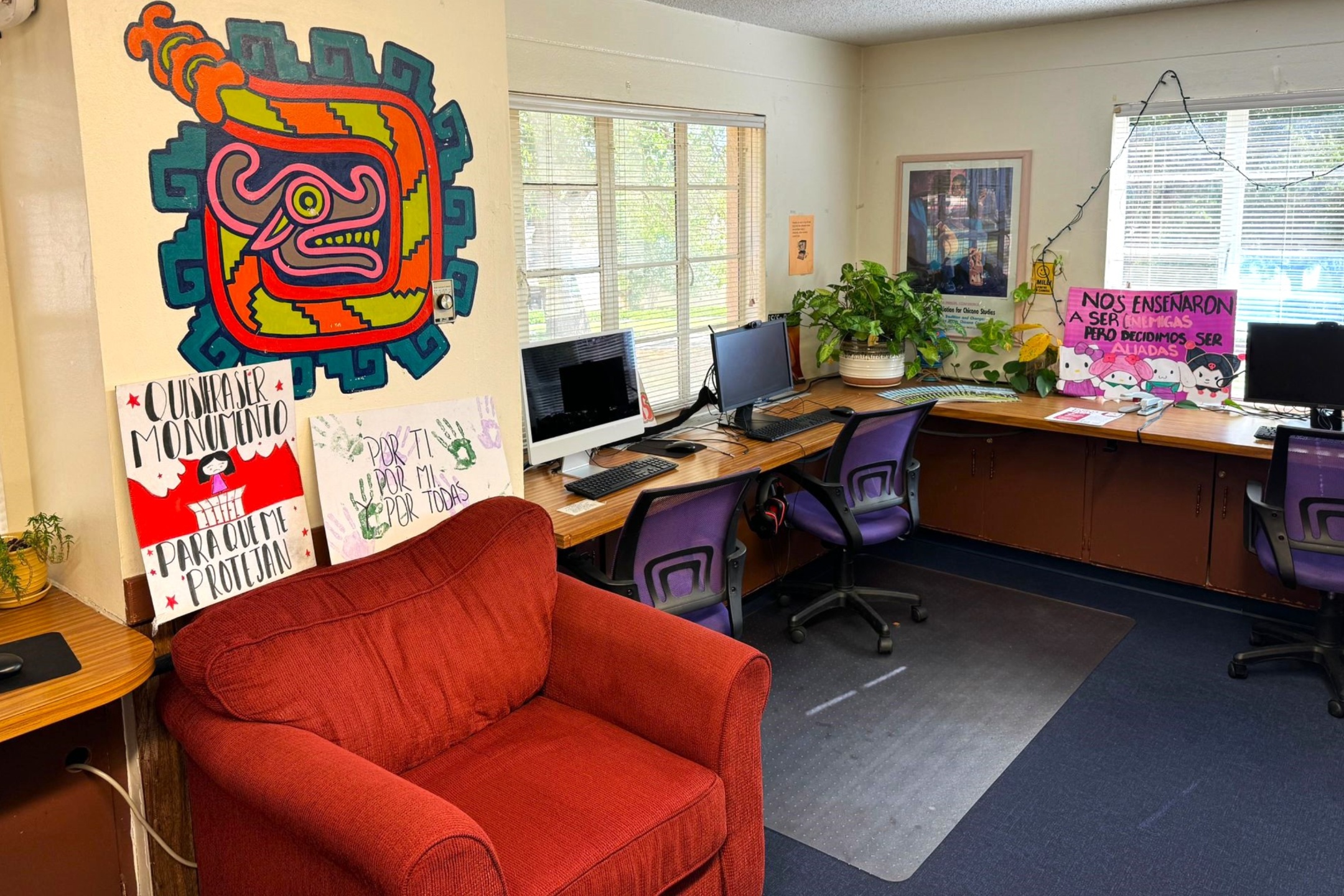 A picture of an the Lab of Latin American Program. The image depicts a small, cozy room that appears to be a workspace or a lounge area. In the foreground on the left, there's a plush red armchair. Behind the armchair and to the right, there’s a corner with a desktop computer setup featuring several desktop computers on a continuous wooden desk against the wall. The desk has three purple office chairs. A poster is mounted above the desk with a colorful and abstract design featuring geometrical patterns and shapes.  On the wall behind the red armchair, two handmade posters rest on the floor against the wall. One has a red section with a figure and the text in black, and the other is white with green handprints and text.  Two windows with blinds allow natural light to filter in, illuminating the room. A large, leafy potted plant in a white pot sits on the desk. On the right side of the image, another handmade poster with pink background text is propped against the wall with illustrations of cartoon characters.