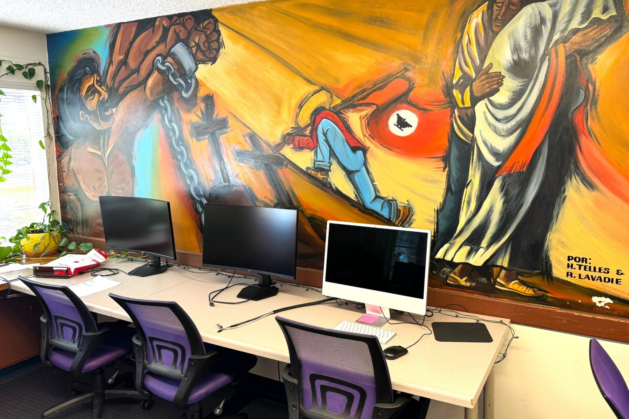 A picture of an the Lab of Latin American Program. The image depicts a mural painted on the wall above a row of desks in a room with multiple computer workstations. The mural is vibrant and spans the entire upper section of the wall, depicting a series of scenes that appear to convey themes of struggle and liberation. The mural is predominantly in shades of yellow, orange, blue, and red colors. The left side shows a muscular figure breaking free from chains, with intense facial expressions and vibrant colors highlighting the action. Towards the center, a figure in a red jacket and blue pants kneels on the ground as if in a moment of effort or reflection, with a large, red, circular emblem featuring a black silhouette of an eagle behind it. On the right, a figure draped in a white and orange garment stands with a solemn expression. The mural is signed in the bottom right corner by "H. Telles & R. Lavadie." Below the mural, there are four workstations with computer monitors, keyboards, and chairs. To the left, there's a window with a curtain partially drawn up and a trail of green hanging plants on the windowsill and wall.