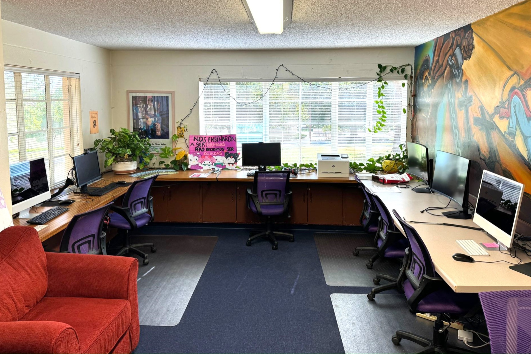 A picture of an the Lab of Latin American Program. The image depicts an office room furnished with several desks, chairs, and computer stations. The layout forms a U-shape along three walls. The left side of the room has two desks with desktop computers and large monitors, complemented by purple mesh office chairs on wheels. Adjacent to these desks is a large potted plant and a framed picture on the wall.  The back wall features a wide window that spans almost the entire length of the room, allowing ample natural light to enter. Below the window is a long desk with an assortment of items on it. The items include a computer monitor, another large potted plant, various papers, and a pink poster with text and illustrations. There is also a printer located near the right end of this desk. Decorative string lights and cascading green plants enhance the window's appearance.  The right wall has a mural with vibrant colors depicting various figures in motion. Below the mural are several computer workstations, similarly equipped with desktop computers and purple office chairs. An empty desk is nestled at the room's center.  A red fabric couch is positioned in the foreground on the left side of the room, adding a cozy element to the otherwise utilitarian setup. The floor is carpeted in dark blue with clear plastic floor mats placed under the chairs to protect the carpet.