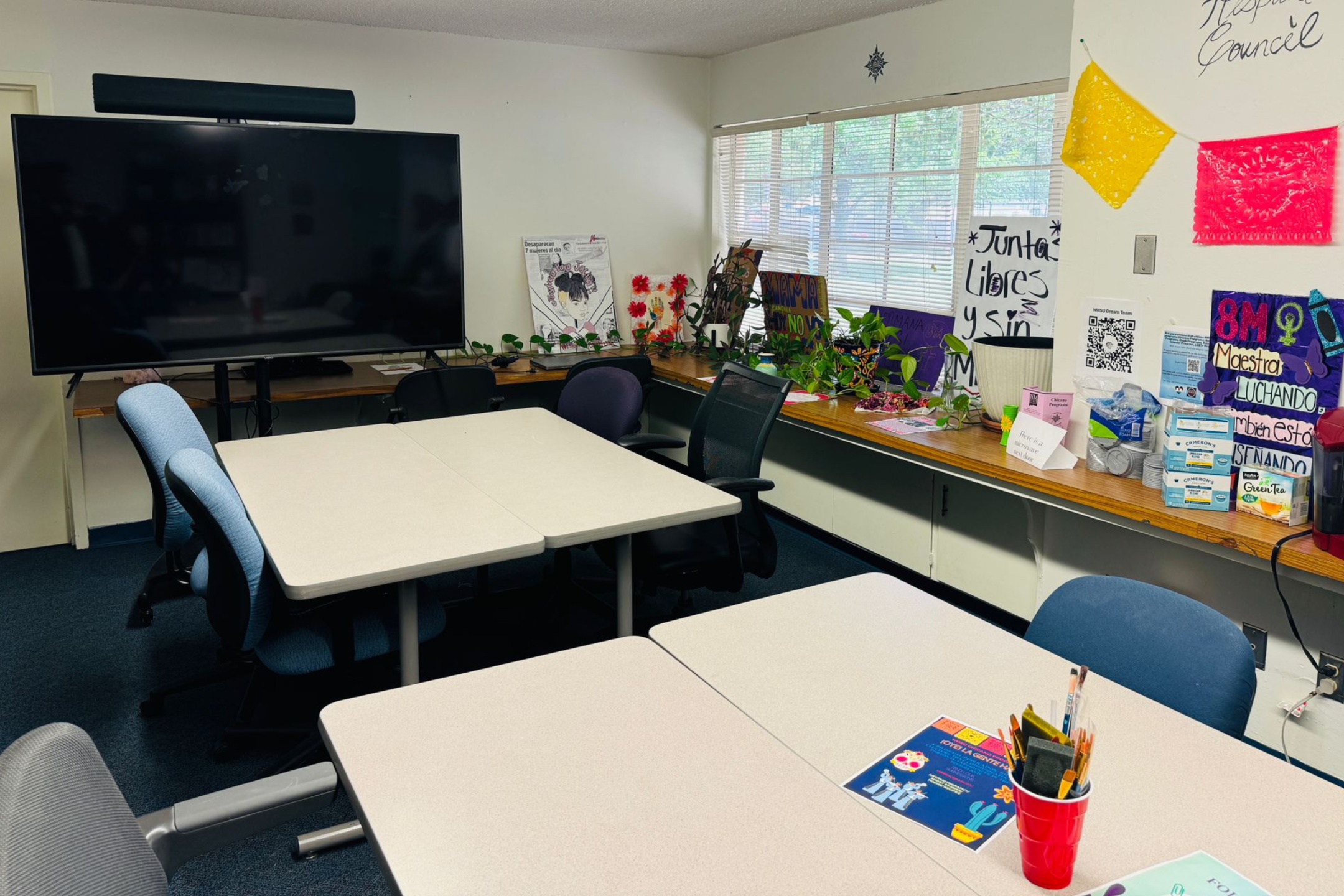 A picture of an the Lab of Latin American Program. The image depicts an interior of a classroom or meeting room equipped with a variety of furniture and decorations. In the foreground, there are two light-colored rectangular tables placed together with office chairs around them. On the closest table are two flyers and a red cup filled with markers and office supplies. In the background, there is a wall-mounted large flat-screen television with a soundbar above it. Next to the television is a wooden shelf running along the wall below a set of windows with closed blinds. The shelf holds various colorful decorations and plants. On the right side of the image, several posters and signs with drawings and text in both Spanish and English are attached to the wall and shelf. Yellow and pink papel picado decorations are hanging on the wall near the posters. Office supplies and a stack of tea boxes are also visible on the shelf.