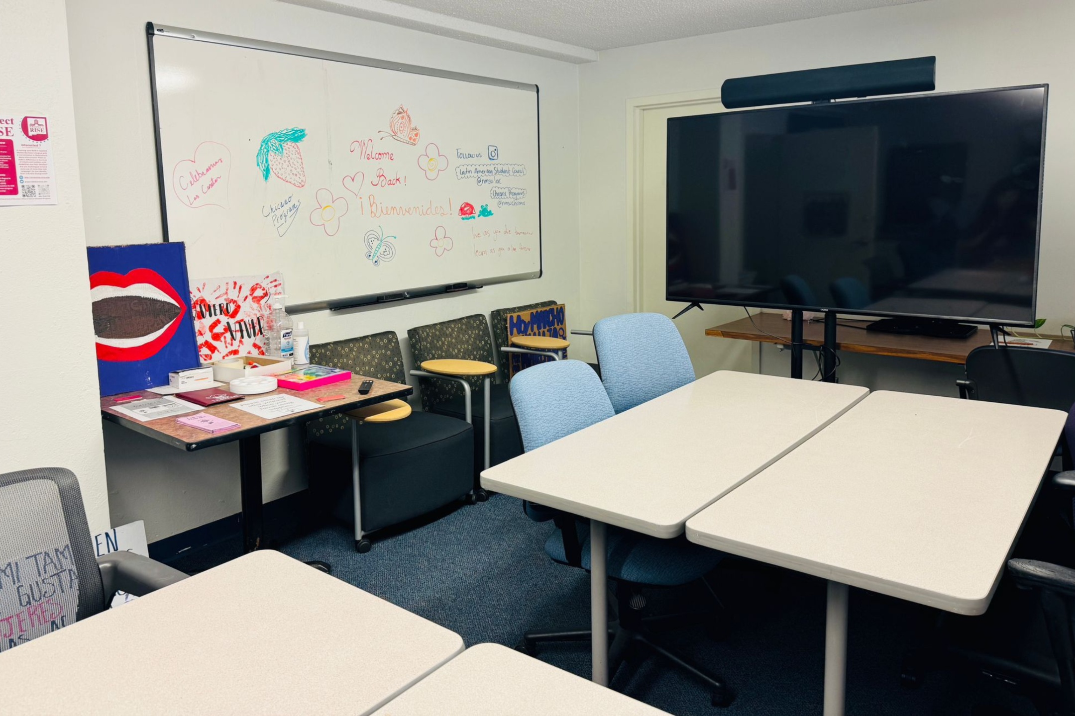 A picture of an the Lab of Latin American Program. The image showcases a small office or classroom space. The room contains two off-white rectangular tables pushed together, surrounded by several chairs with light blue and dark gray fabric upholstery. Against the wall to the right is a large flat-screen TV on a black stand. To the left, there is a whiteboard filled with colorful drawings and text, including hearts, flowers, a strawberry, and welcoming messages. Below the whiteboard, there is a black table with various items like hand sanitizer, bins, flyers, and artwork, including a prominent painting of a pair of red lips. The room has a blue carpeted floor and white walls. In the back left corner, there are a few more chairs with attached writing surfaces stacked near the table.