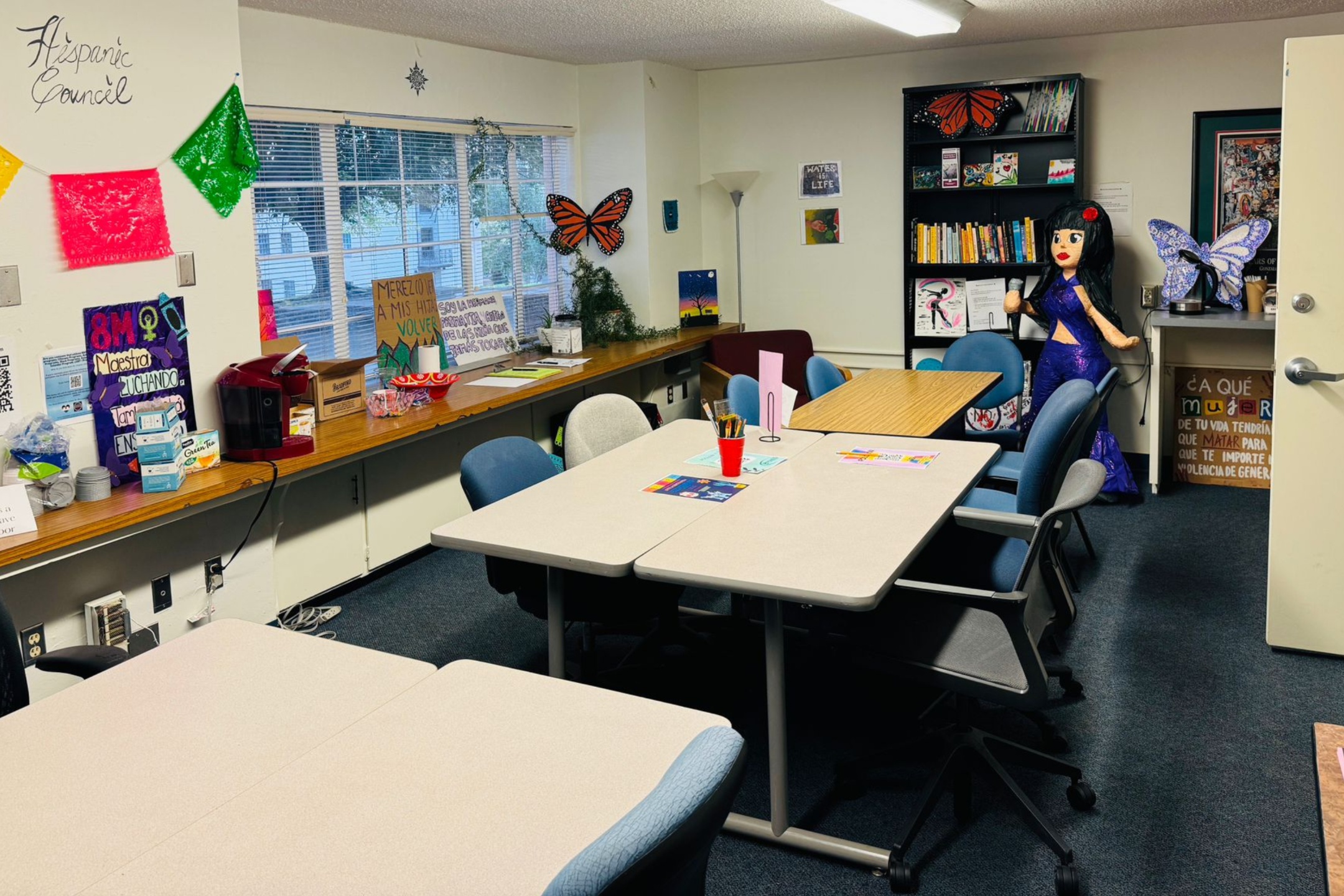 A picture of the study room of Latin American Program. The image depicts a study or meeting room with various decorations and furnishings. The room has a dark carpeted floor and off-white walls. There are two rectangular beige tables placed in the center, surrounded by multiple cushioned chairs in different colors (blue, gray, and black).  In the left corner of the room, a long wooden counter runs against the wall below a wide window with blinds partially open, showing trees outside. On the counter, there is a red coffee machine, snacks, and a variety of supplies including face masks in boxes, a tissue box, and a sign. Colorful paper banners are strung across the top of the window.  On the right side, a black bookshelf filled with books and decorations leans against the wall. Next to the bookshelf stands a large figure of a woman in a purple dress, likely a piñata, with butterfly wings behind it. Several posters and colorful signs with text and images are attached to the walls and surfaces throughout the room.  Text Section:  "Hispanic Council" (on the wall above the paper banners) "8M Maestras Luchando" (on a poster on the counter) "Merezco a mis hijas volver a ver" (on a poster) "Maybe Life" (on a piece of art on the wall) "CA QUE MUDAR DE TU VIDA TENDRIAS QUE MATAR PARA QUE TE IMPORTE VIOLENCIA DE GENERO" (on a poster behind the large figure)