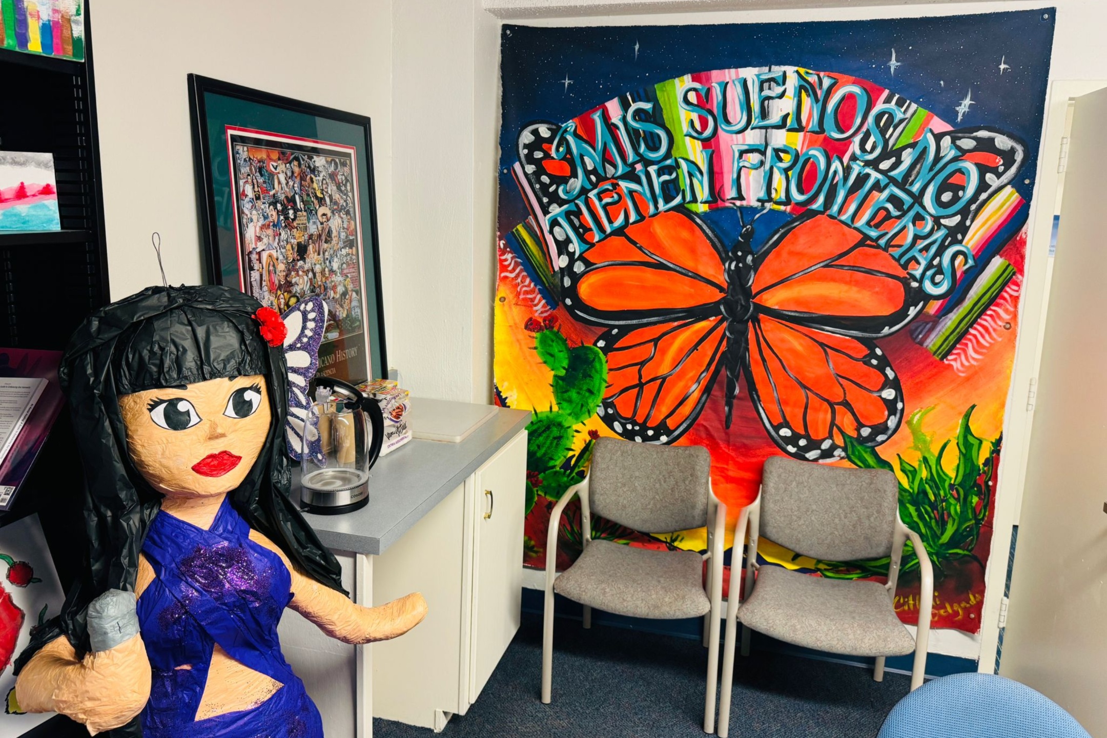 A picture of an the Lab of Latin American Program. The image depicts an indoor setting with various decorative and functional elements. Dominating the right side of the image is a vibrant mural on the wall featuring a large orange butterfly with black and white accents. Above the butterfly, colorful, stylized text reads, "MIS SUEÑOS NO TIENEN FRONTERAS." Below the mural are two beige, cushioned chairs placed against the wall.  On the left side, there is a corner with a countertop in gray and a white cabinet underneath. On top of the counter is a clear, electric kettle and a boxed item beside it.  In the foreground, left side of the image, stands a large doll or piñata of a woman with long black hair, a red flower on her head, and large, expressive eyes. She is dressed in a purple outfit and holds a gray microphone.  Above the counter is a framed, colorful collage image related to Chicano history. On the far left, a portion of a black shelf is visible, containing what appears to be paper and colorful items.